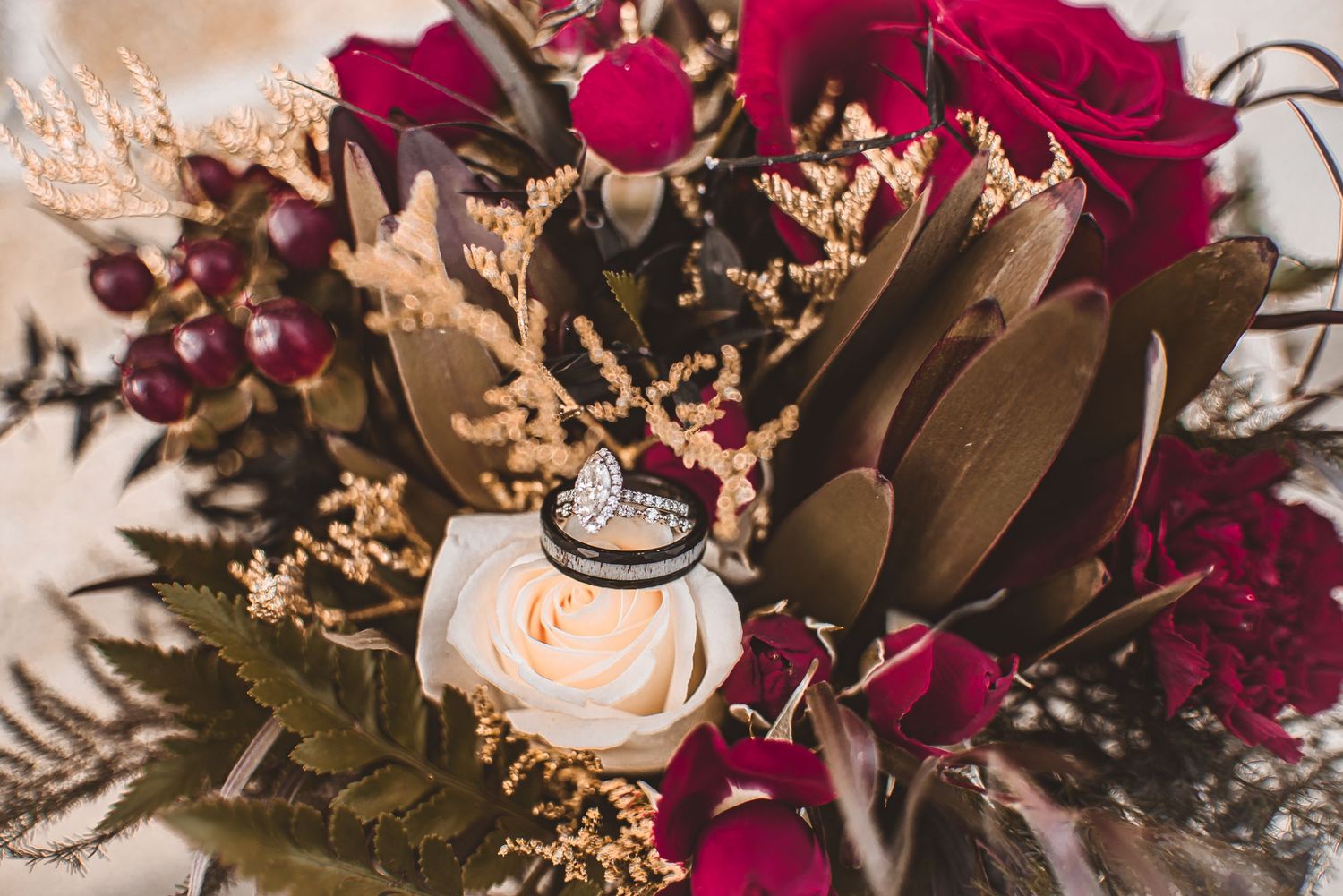 Wedding rings nestled among deep red roses and dried flowers in a moody, romantic arrangement.