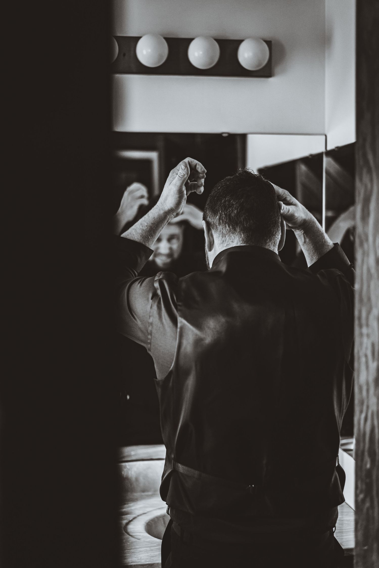 Back view of someone adjusting hair in a bathroom mirror with vintage-style lighting.