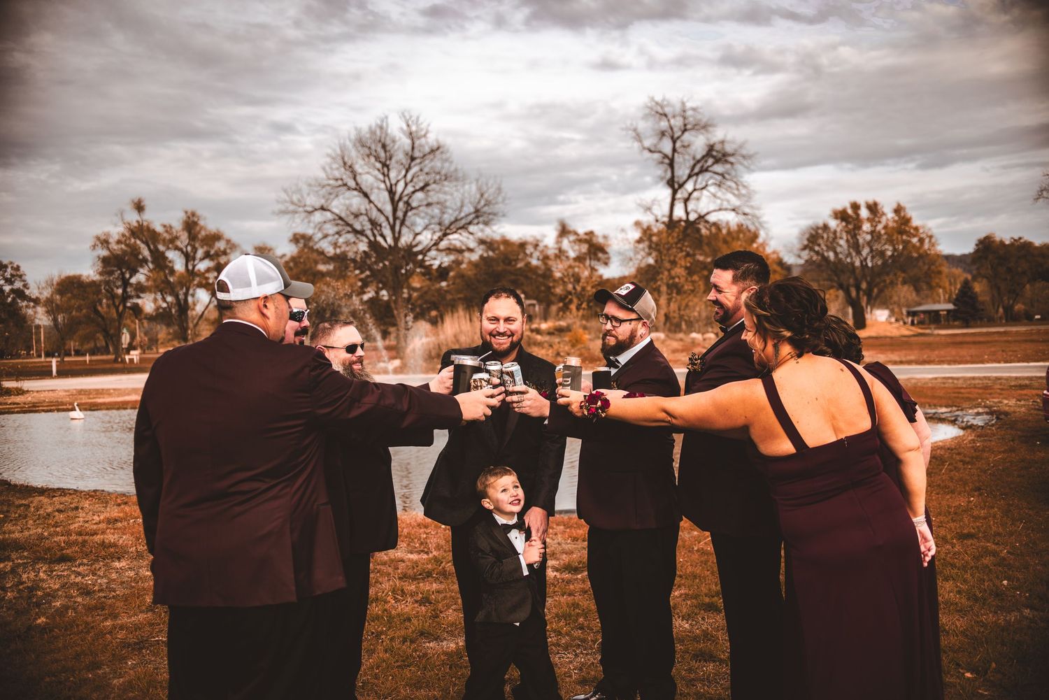 Wedding party and young ring bearer toast together outdoors under cloudy autumn sky.