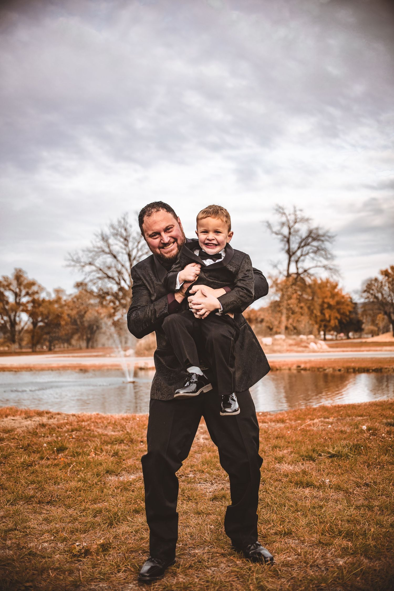 Father and son share a playful moment by a lake on an autumn day.