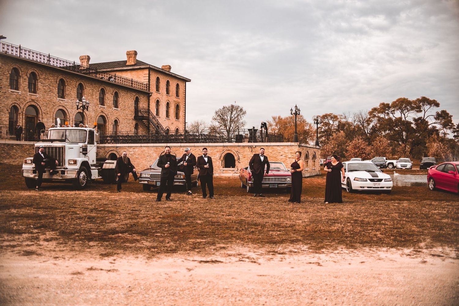 Guests gather outside a historic brick building with cars parked on dirt ground on an overcast day.