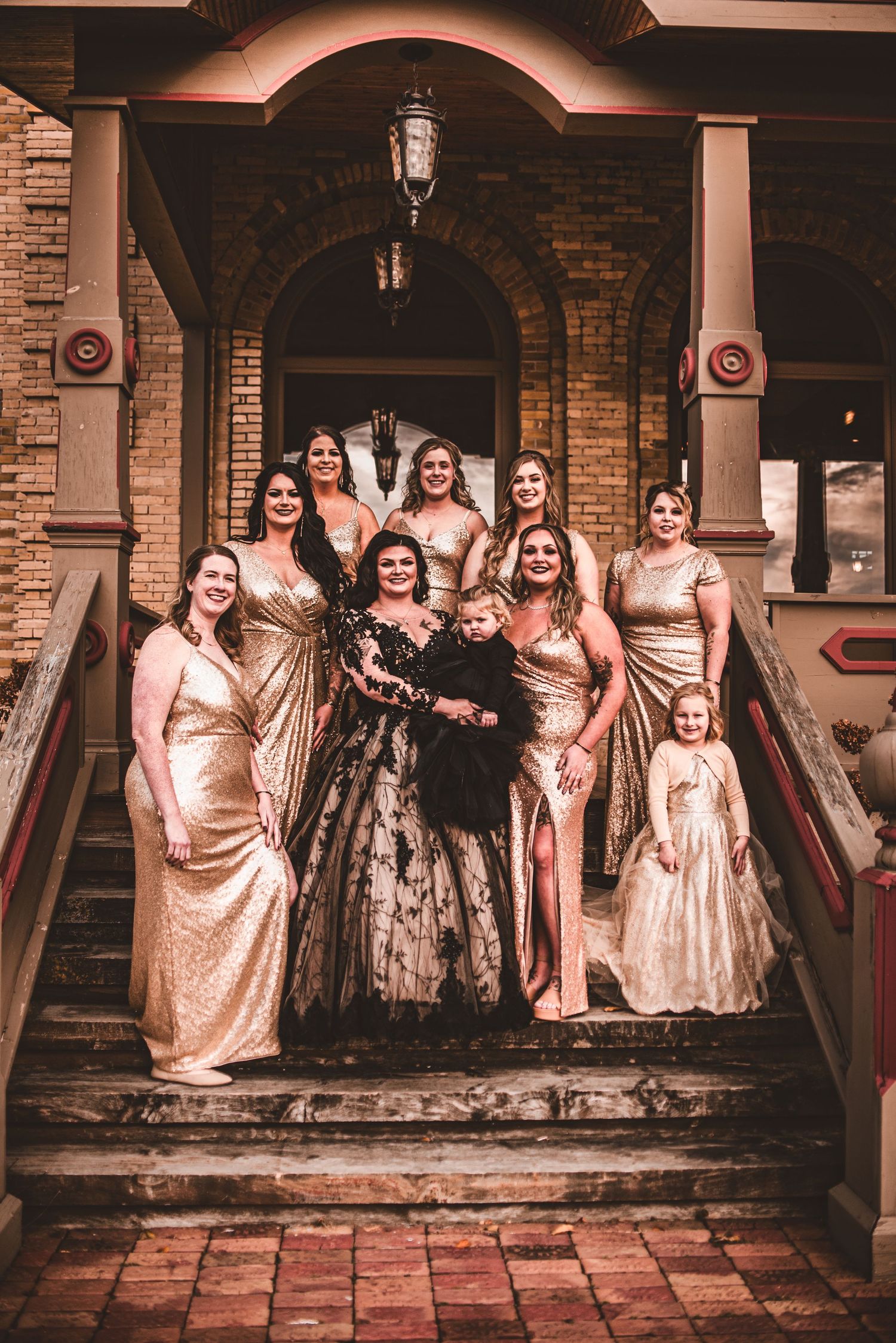 Wedding party in gold dresses poses on brick steps of ornate historic building entrance.