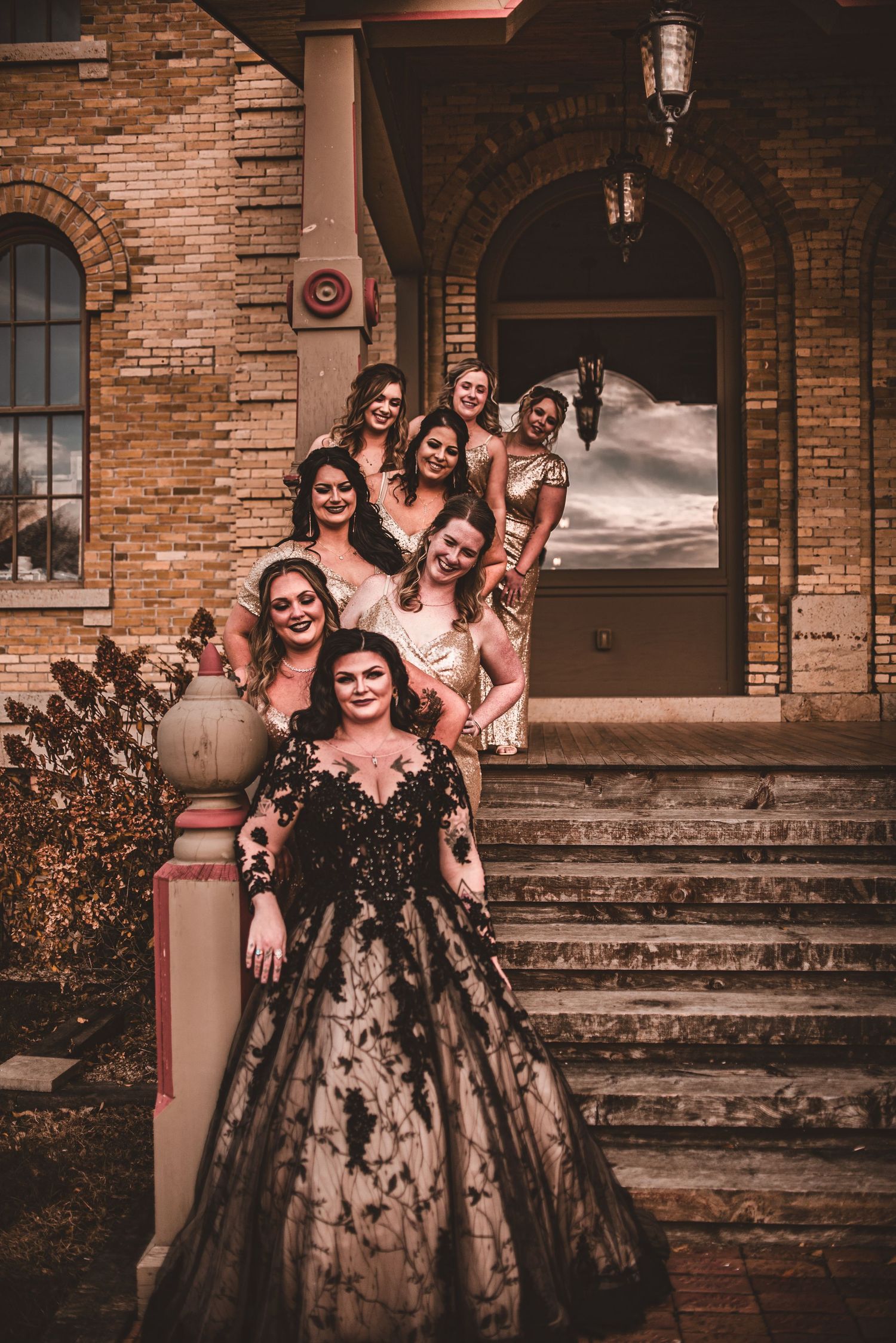 Bridal party in rose gold dresses and black lace gown stands on stone steps of vintage brick building.