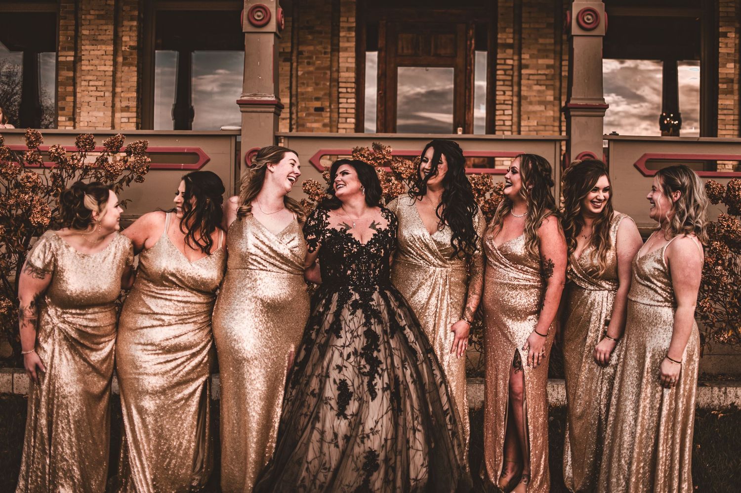 Wedding party wearing metallic gold gowns and black lace dress poses in front of brick archways.