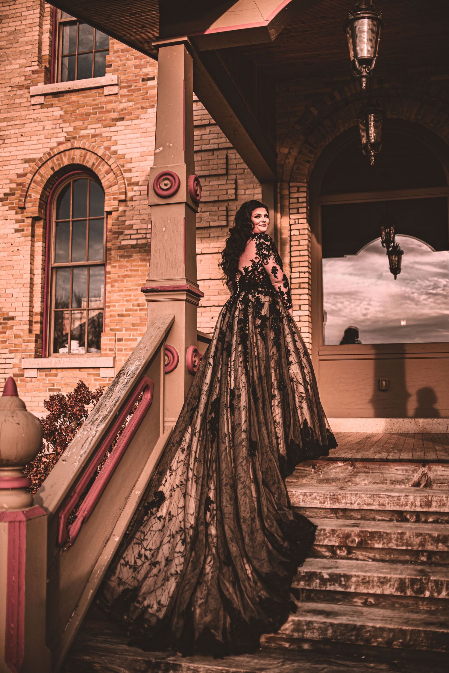 Dramatic black lace wedding gown with long train photographed on vintage brick building steps.