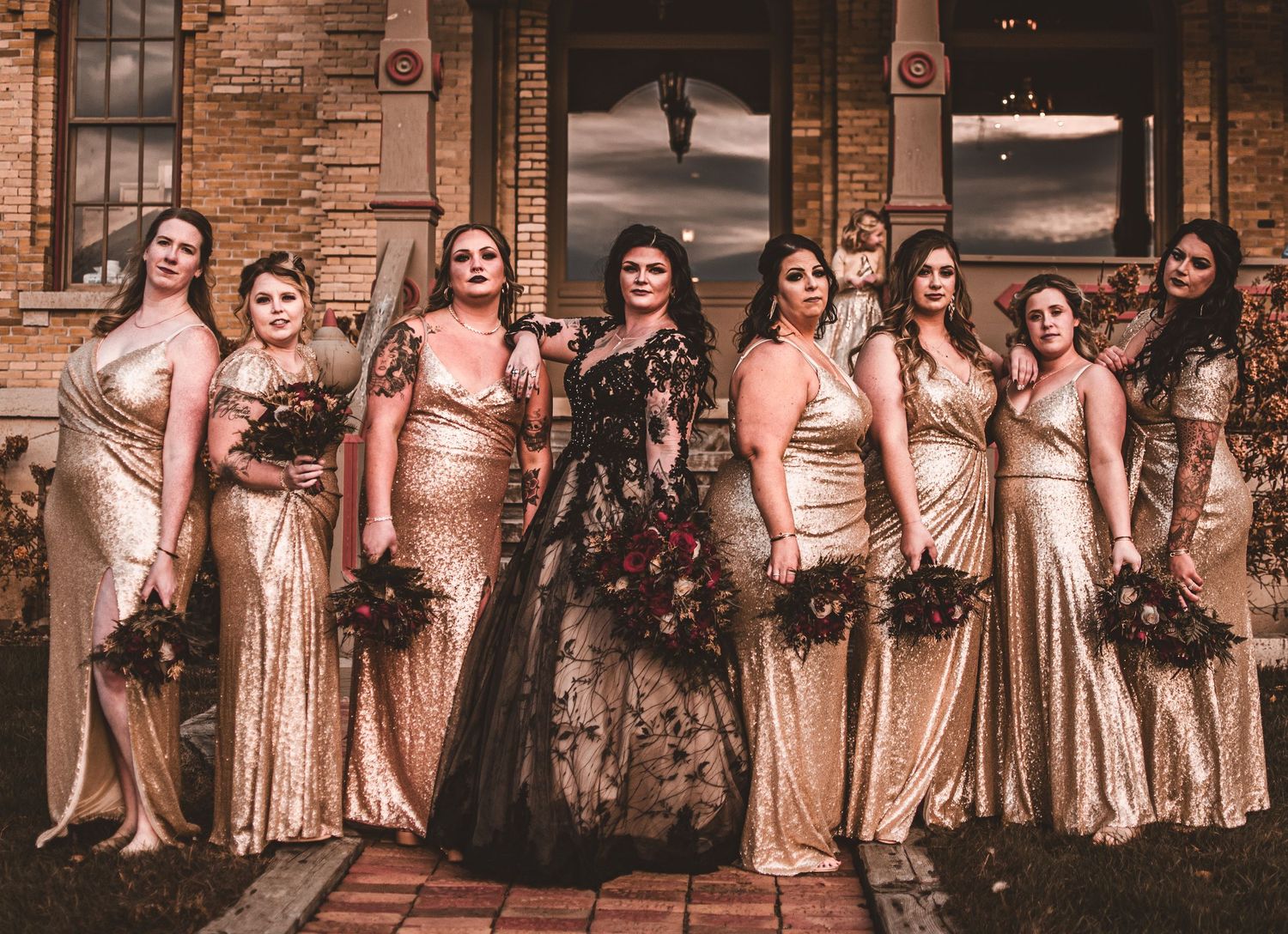 Wedding party in champagne dresses poses with bouquets against historic brick architecture.