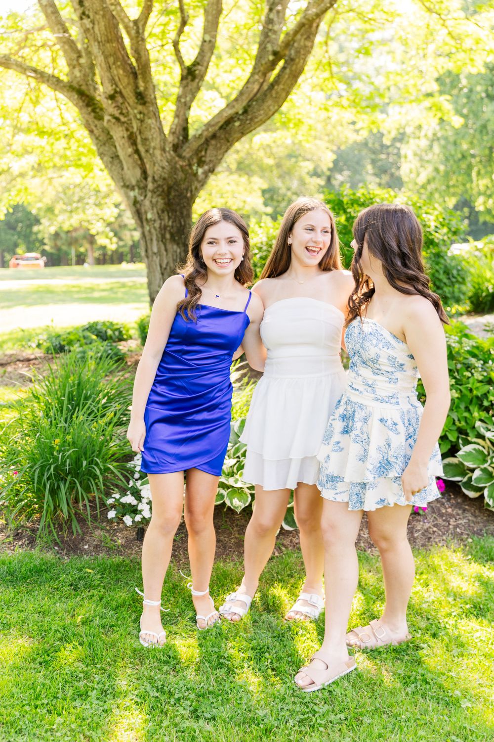 Three friends in summer dresses pose together on a sunny lawn with a large tree in the background.