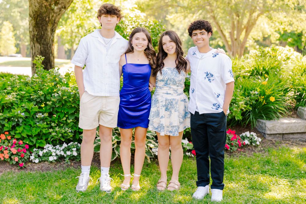 Group photo of four friends dressed in casual attire standing on grass with flowering bushes behind them.