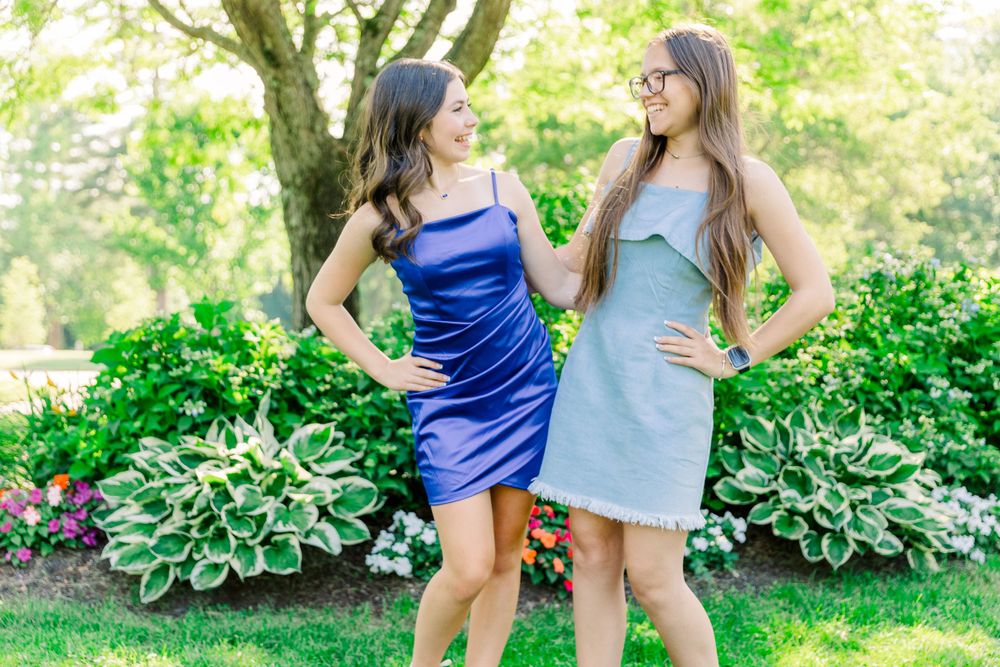 Two friends in blue and grey dresses pose together in a blooming summer garden.