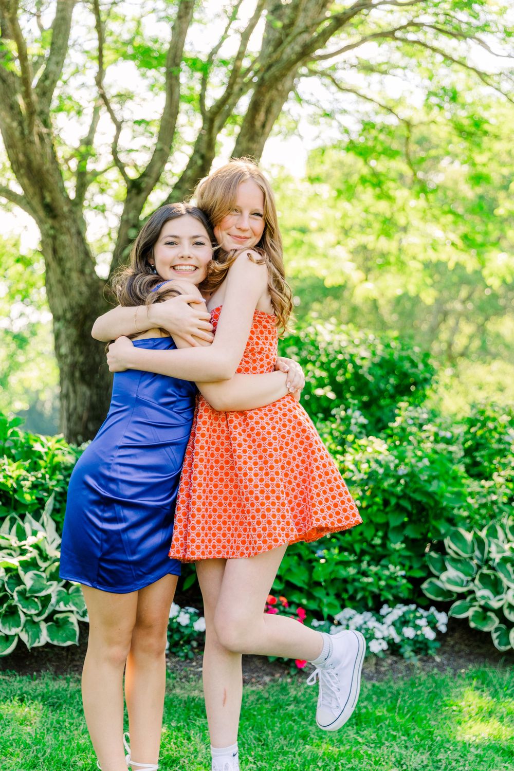 Two friends share a joyful hug outdoors in front of a leafy tree and garden.