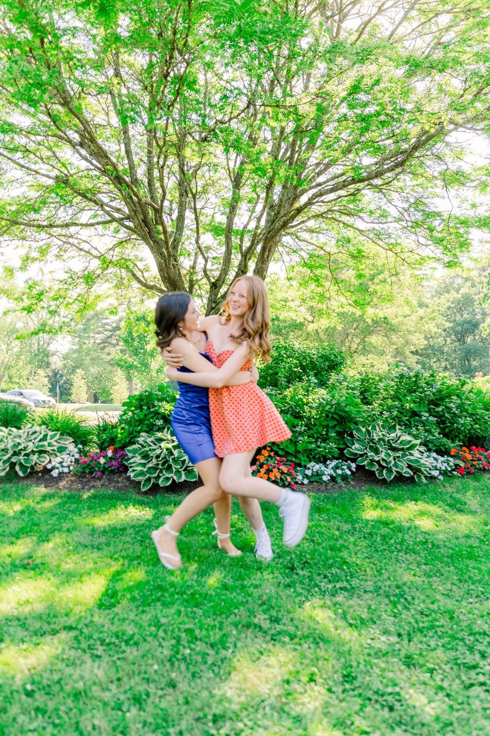 Friends in blue and orange dresses embrace while spinning on a sunny lawn.