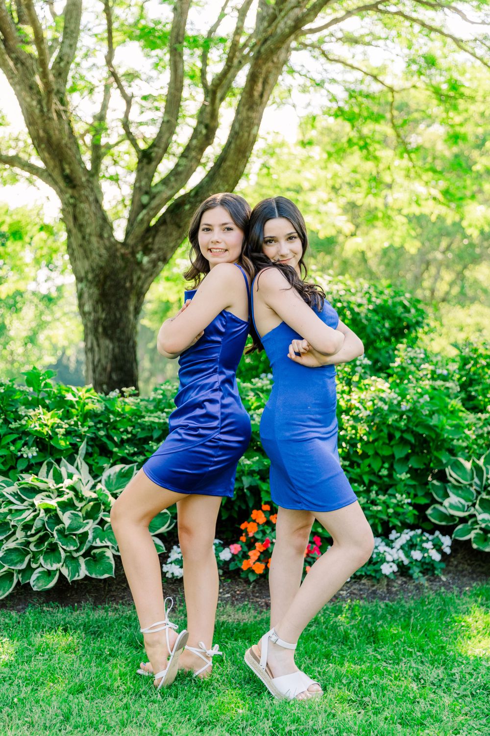 Two friends in blue dresses strike back-to-back poses in a flowering garden.