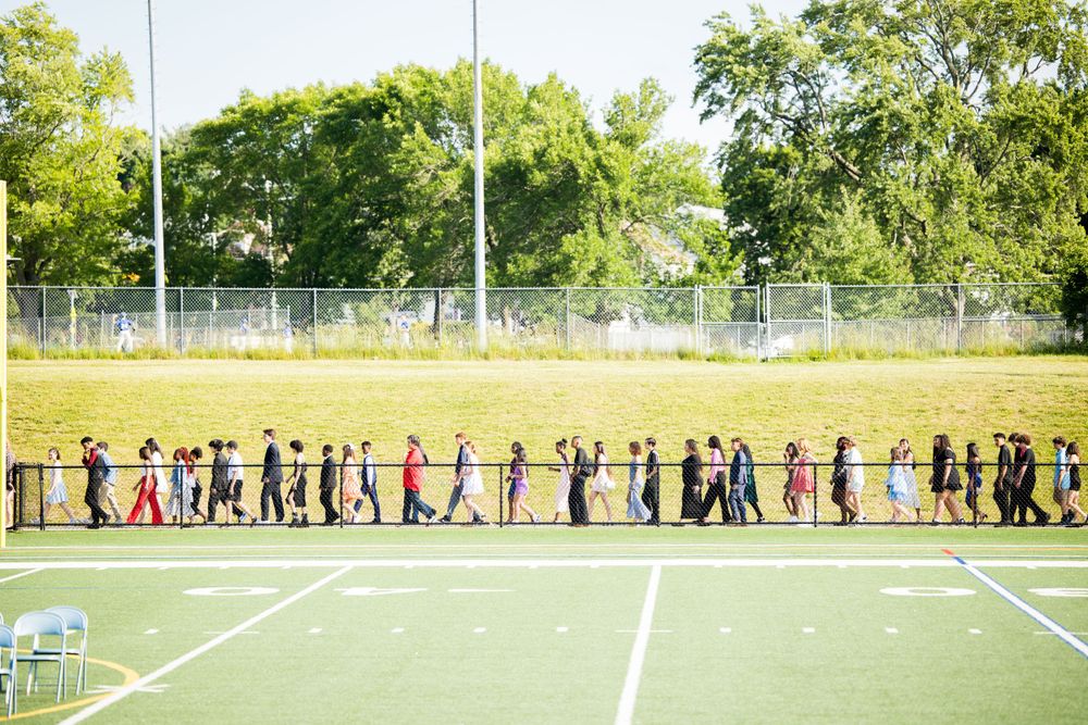 Long line of graduates in formal attire walking across a football field during ceremony.