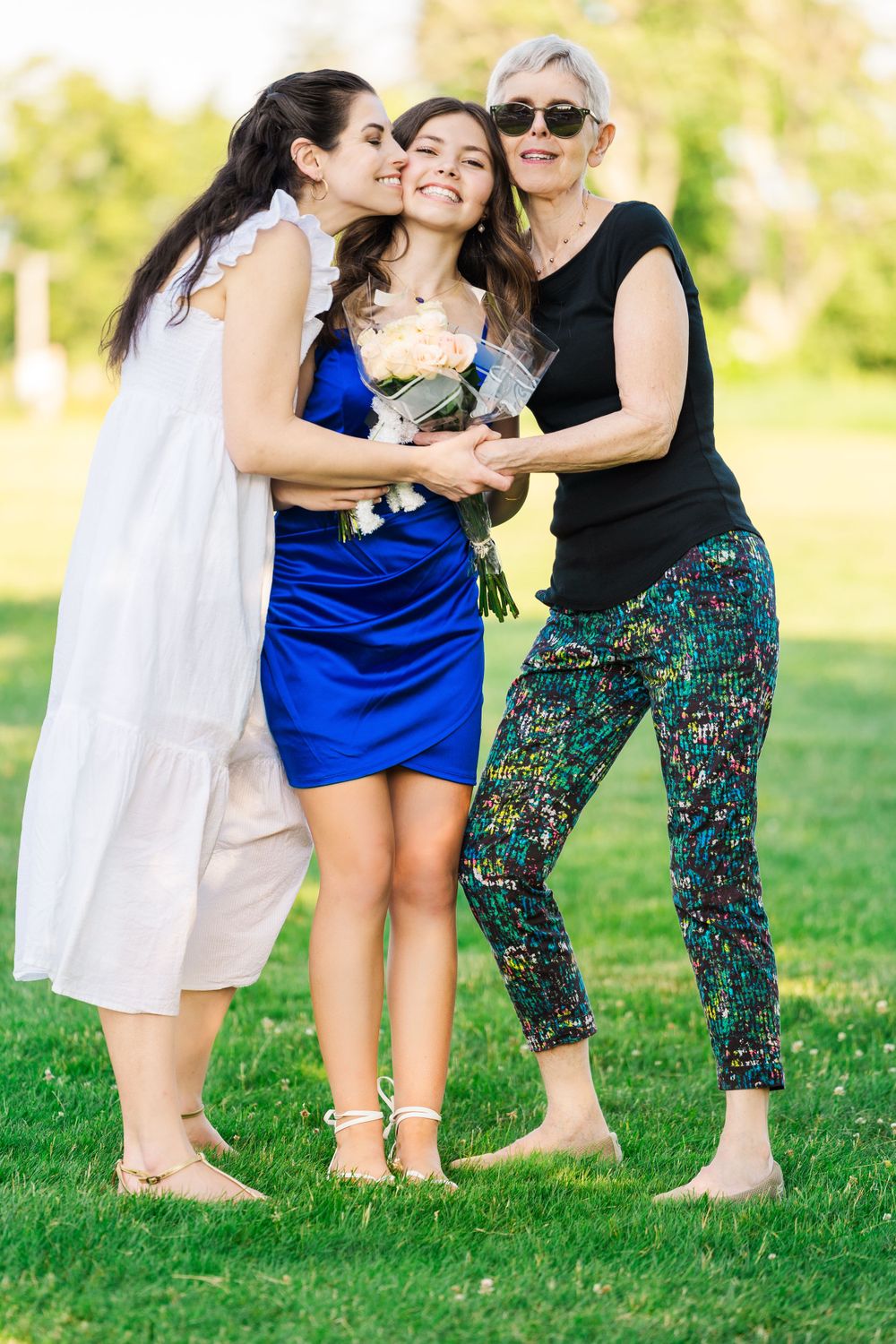 Three people share joyful moment outdoors while holding bouquet on sunny day.