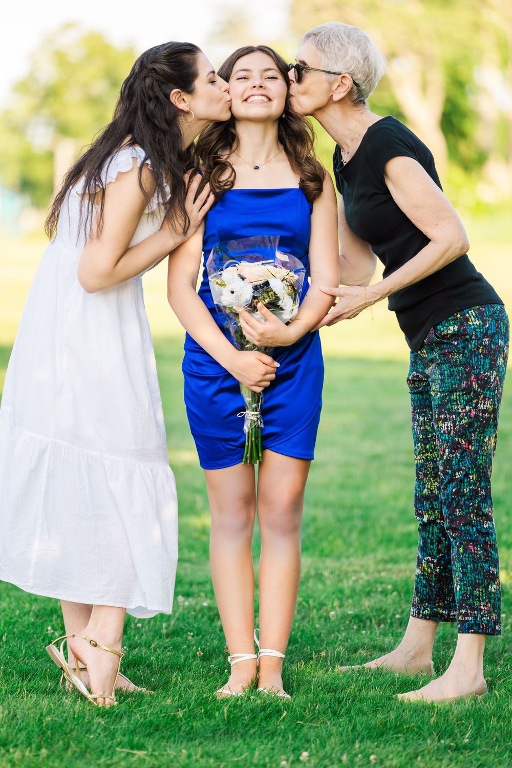 Three generations pose together on grass with flowers, sharing an affectionate moment in summer.