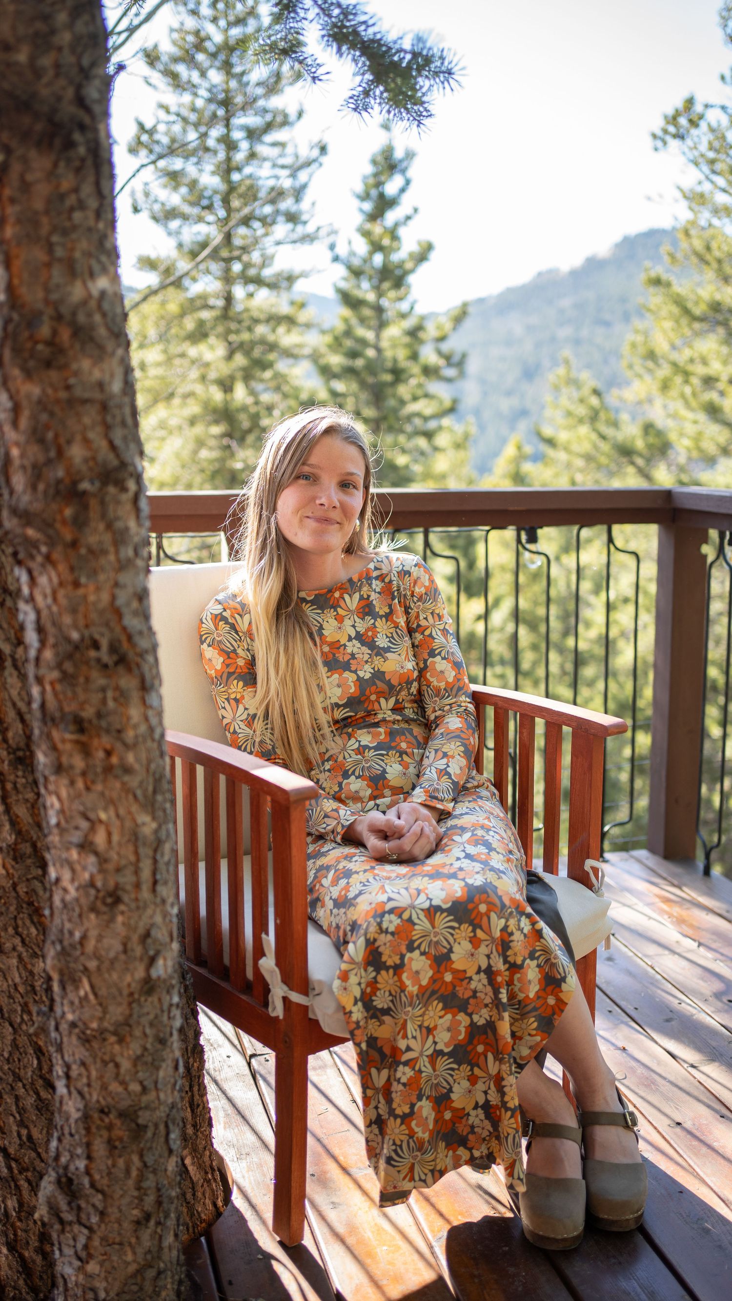 A portrait of a woman in floral dress sits peacefully on wooden deck surrounded by mountain pine trees.