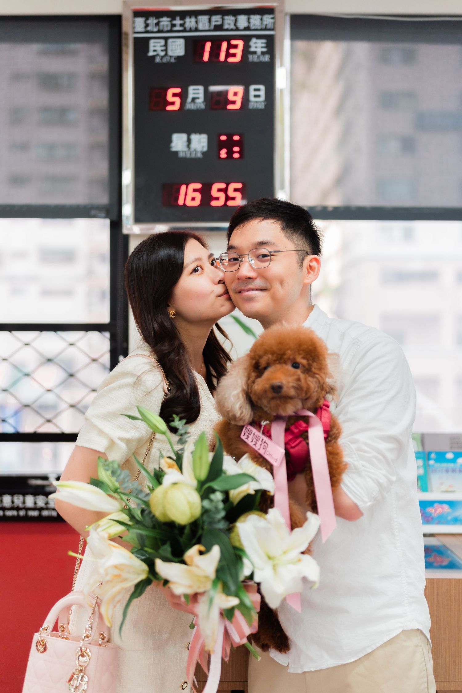Couple poses with small brown dog and bouquet under digital display board.