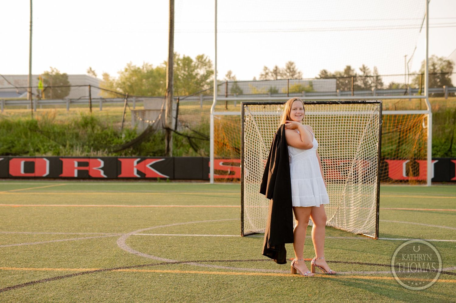Graduate poses in front of lacrosse goal net at sunset wearing white dress and graduation gown.