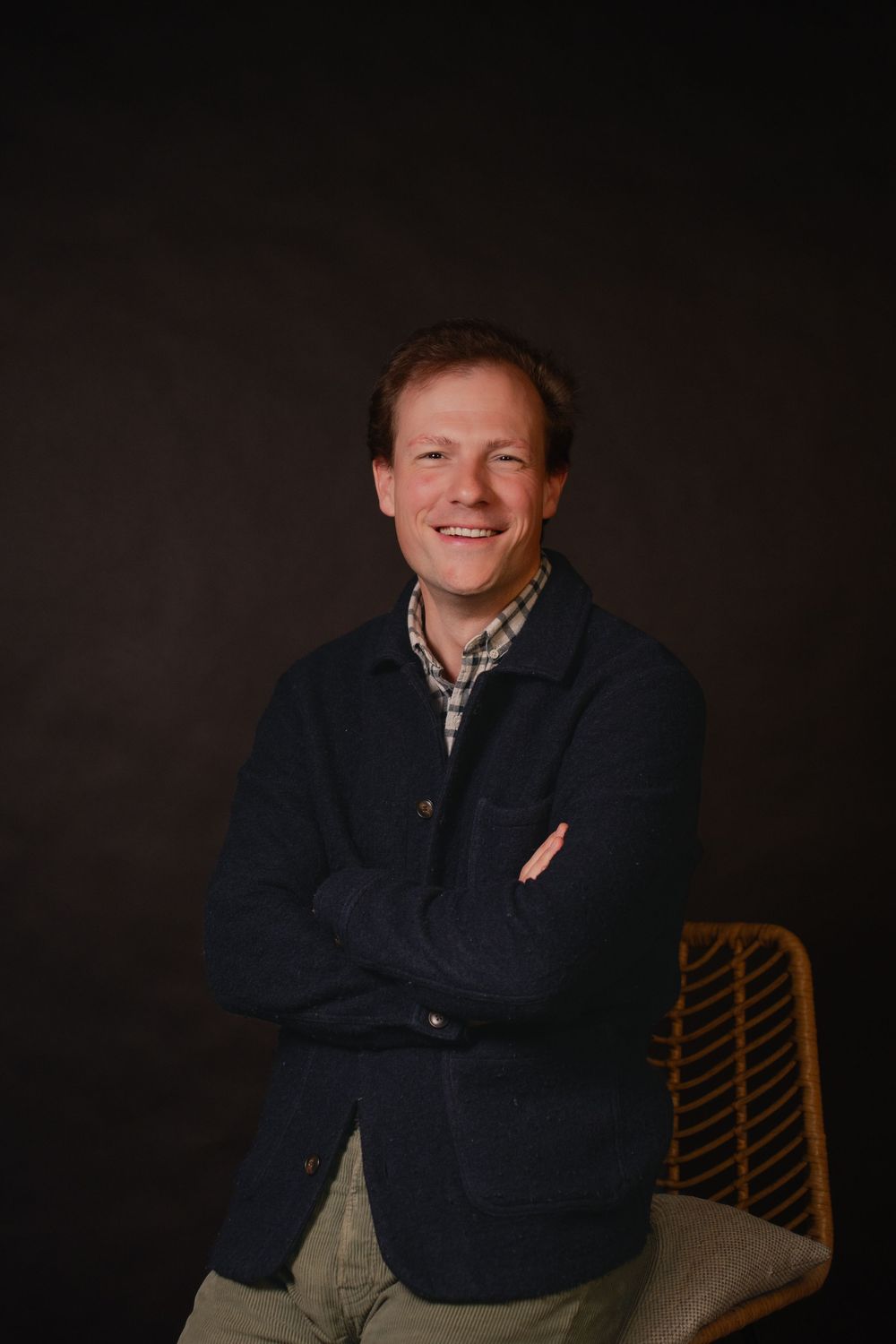 Smiling man with crossed arms in navy jacket and plaid shirt seated on wicker chair against dark background.
