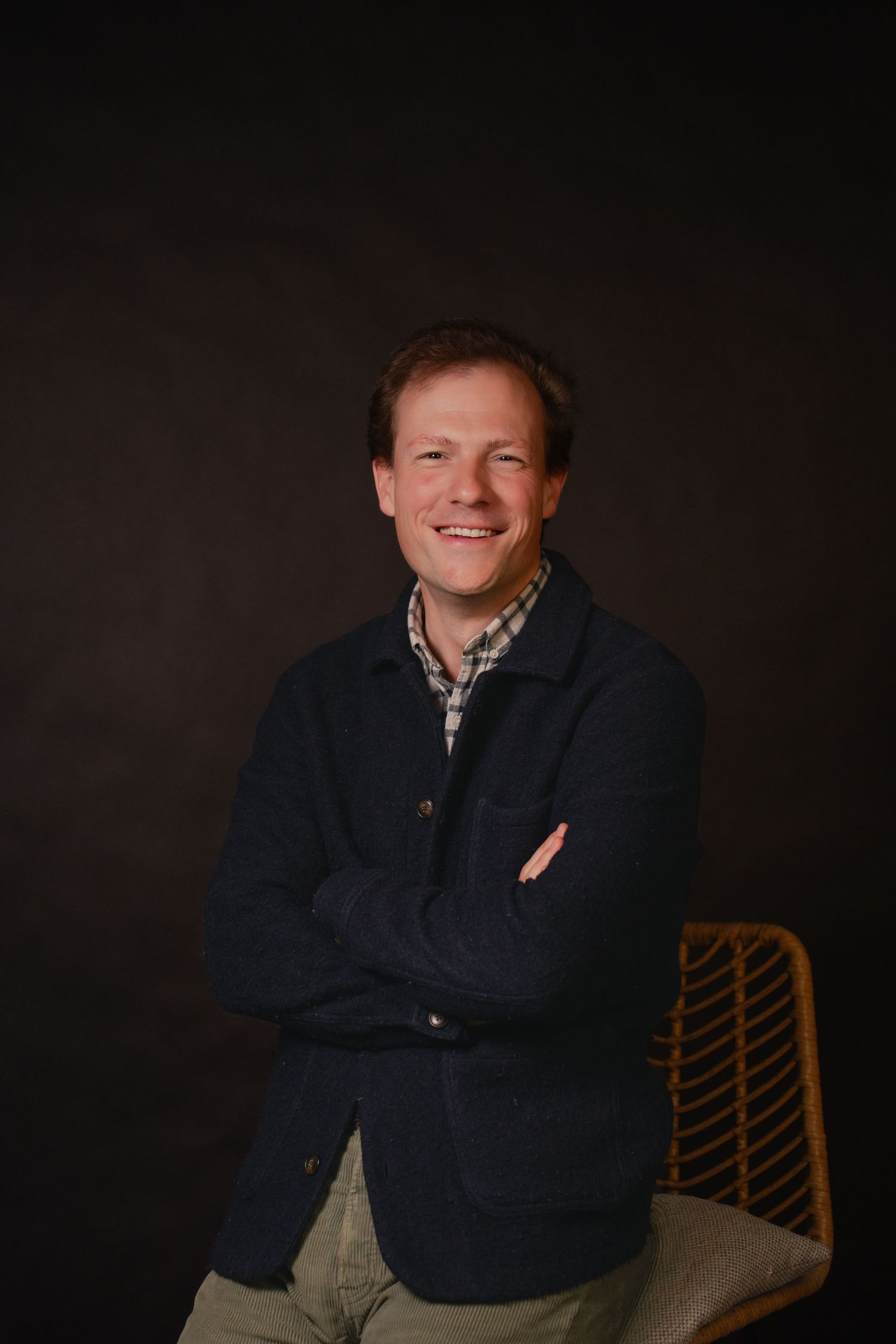 Smiling man with crossed arms in navy jacket and plaid shirt seated on wicker chair against dark background.