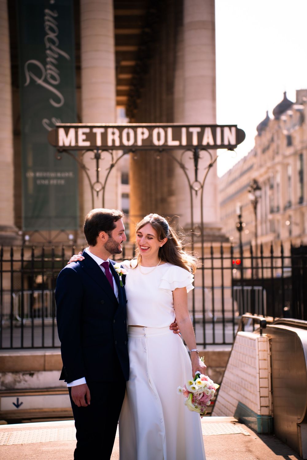 Bride and groom embrace near Paris Metropolitain sign, woman in white dress holding bouquet, man in navy suit.