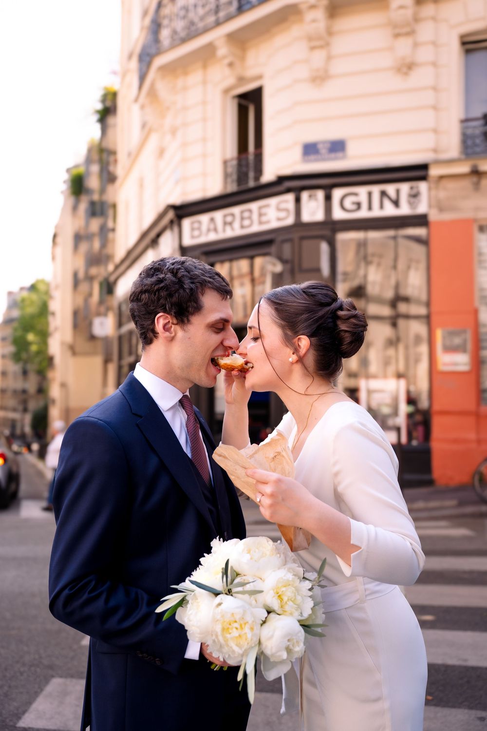 Bride and groom sharing a croissant on a Parisian street, bride holding white peonies bouquet near Barbès Gin sign.