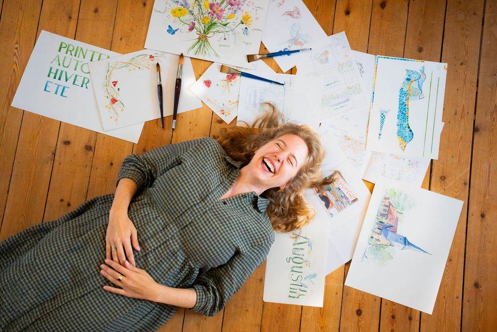 Woman laughing while lying on wooden floor surrounded by watercolor paintings and calligraphy artwork with paintbrushes.