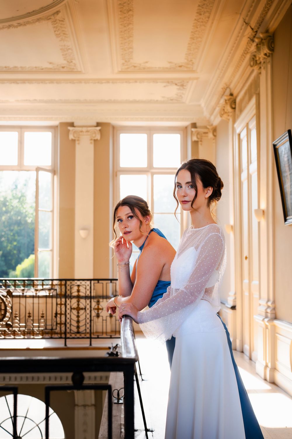 Two elegant women in formal attire pose on a grand balcony in a luxurious venue with ornate decor.