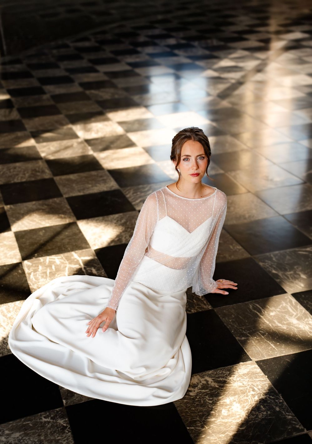 Bride in white gown with sheer long-sleeve top seated gracefully on black and white checkered marble floor.