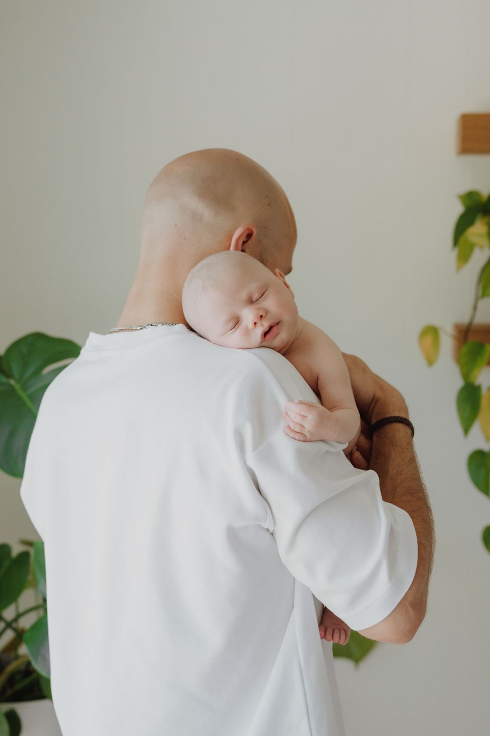 Bald father in white sweatshirt gently holding sleeping newborn baby against his shoulder at home.