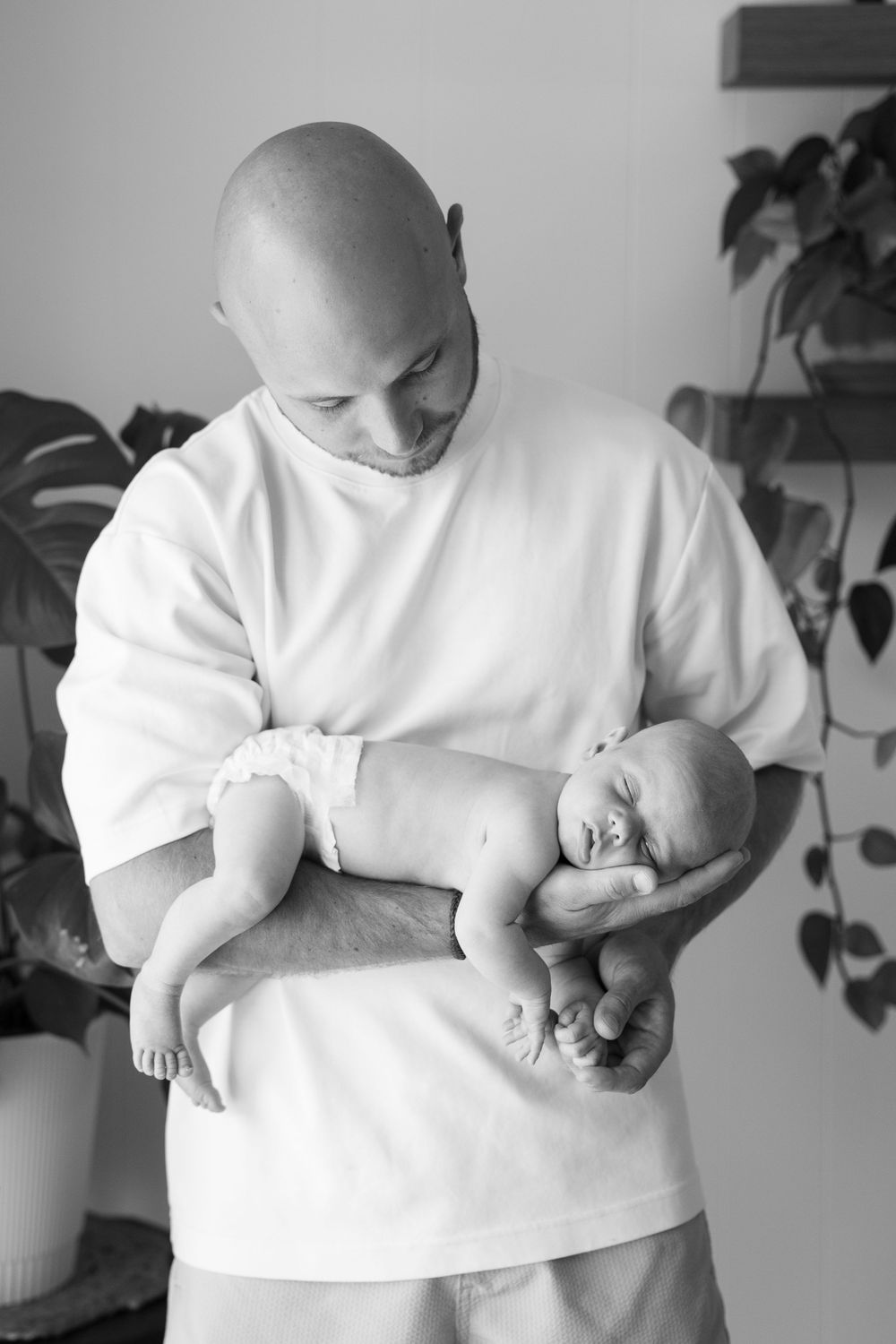 Bald father tenderly holds sleeping newborn baby in arms, black and white portrait with houseplants in background.