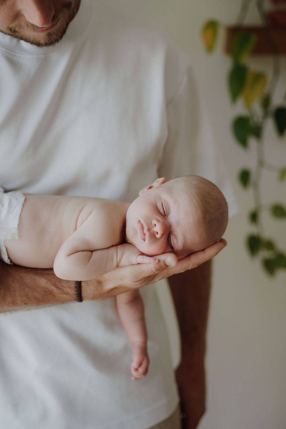 Father in white t-shirt gently cradling sleeping newborn baby in one hand, soft natural light indoor setting.