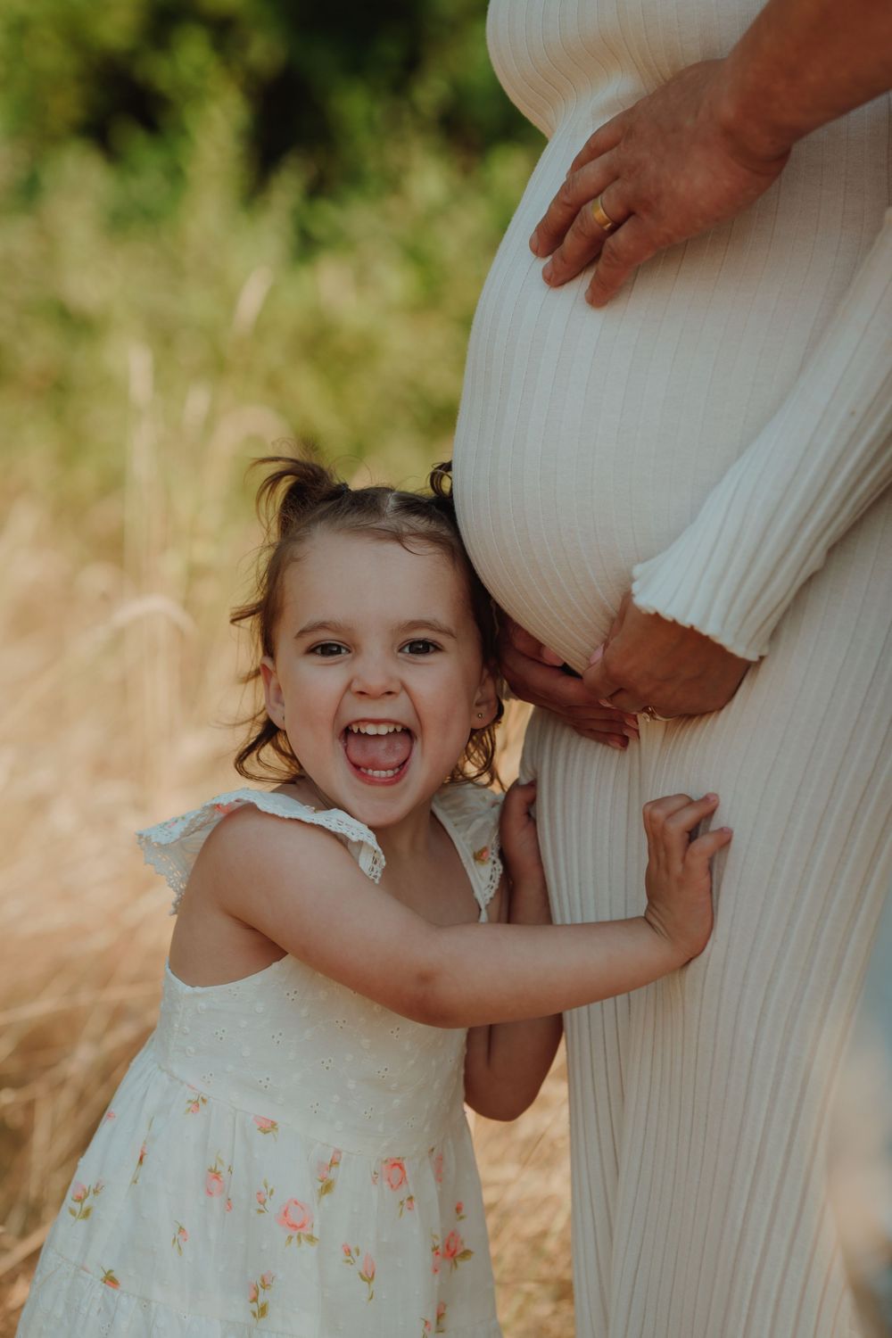 Joyful toddler girl in floral dress hugging her mothers pregnant belly outdoors in golden sunlight. Victoria, BC, Vancouver Island.