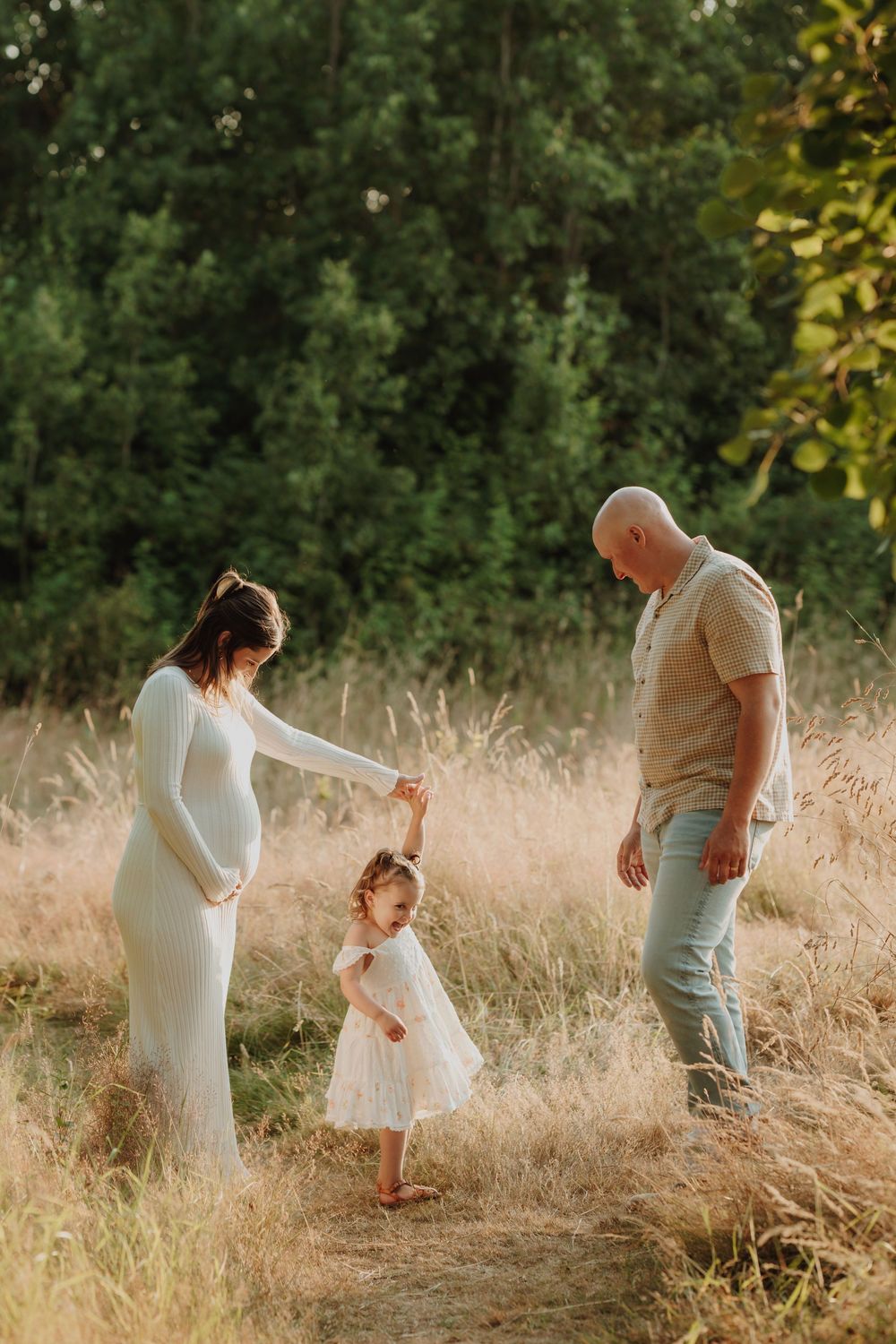 Pregnant mother in white dress holding toddlers hand while bald father watches in a sunlit golden field. Victoria, BC, Vancouver Island.