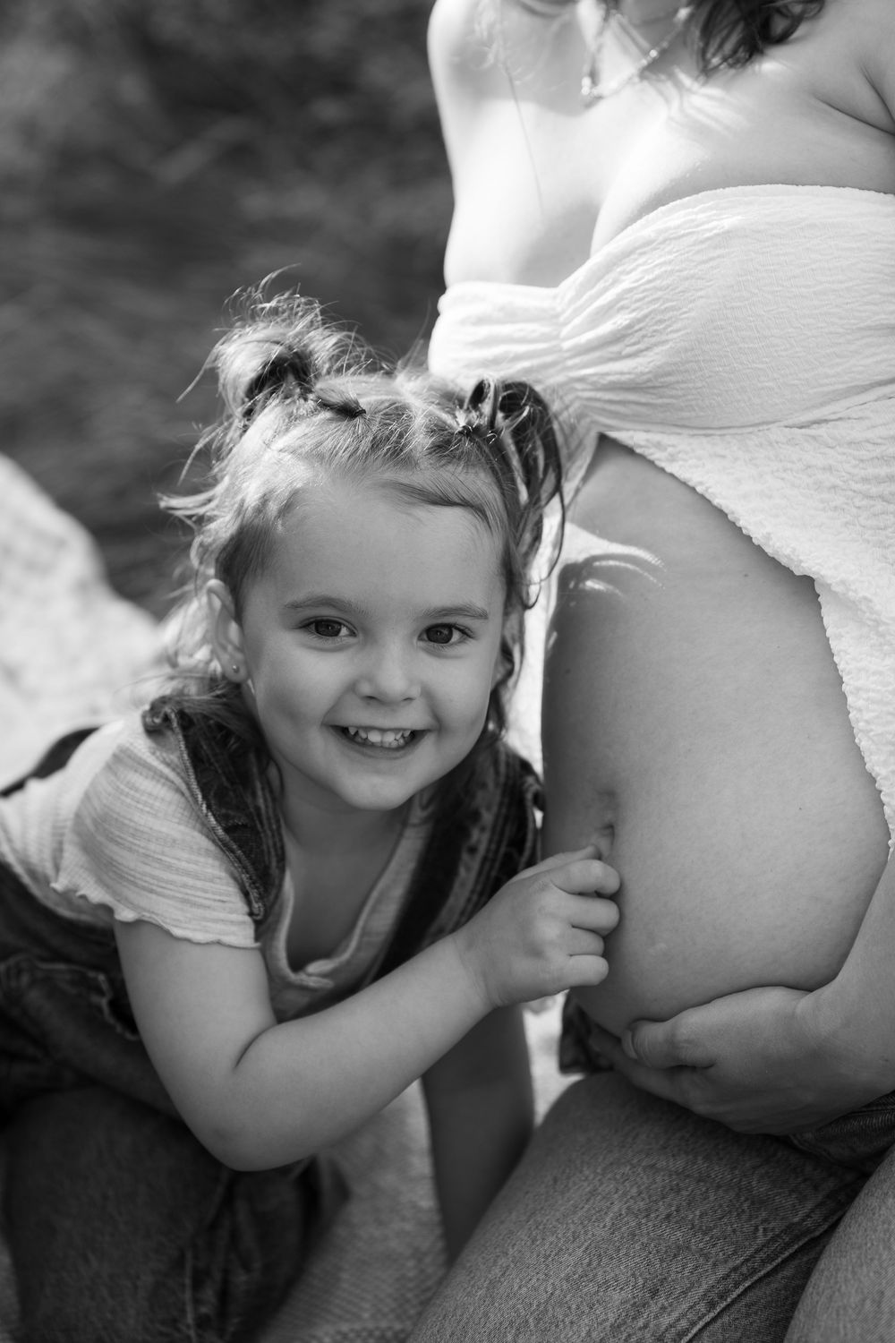Black and white portrait of a smiling young girl leaning against her mothers pregnant belly outdoors. Victoria, BC, Vancouver Island.