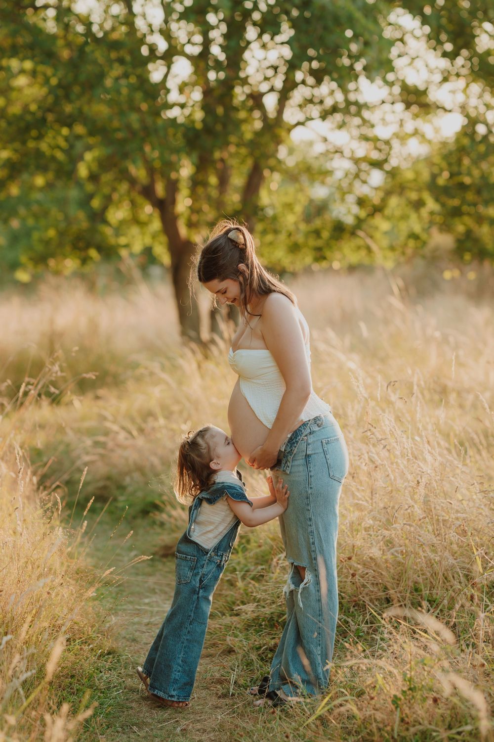 Pregnant mother in white top and jeans sharing a tender moment with her young daughter in a sunlit field. Victoria, BC, Vancouver Island.