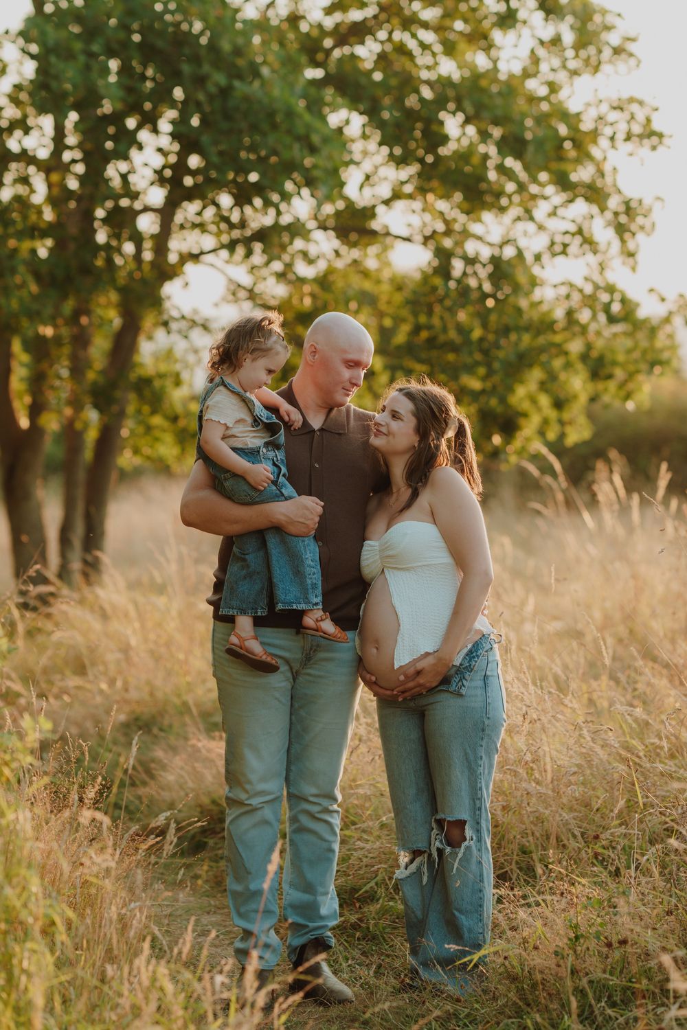 Bald father holding young daughter beside pregnant wife in white top and jeans in a golden sunlit meadow. Victoria, BC, Vancouver Island.