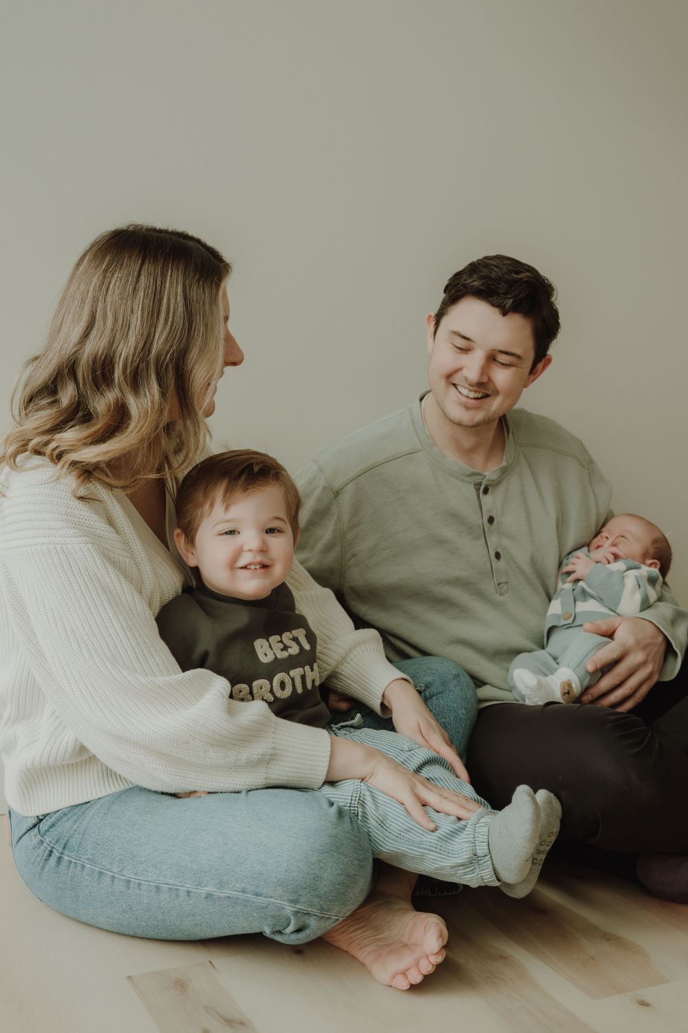 Happy family of four sitting on floor with toddler in best brother bib and newborn baby.