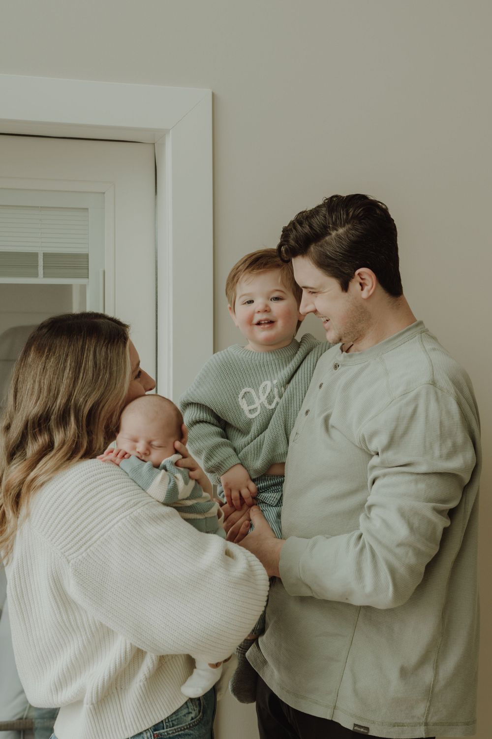 New family of four with newborn and smiling toddler sharing a warm moment in doorway.