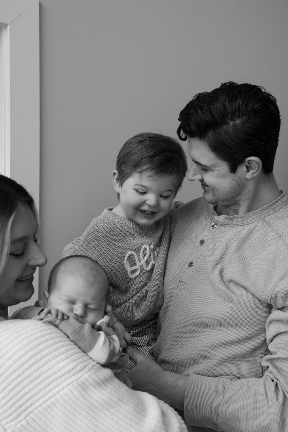 Black and white photo of a family of four admiring their sleeping newborn baby together.