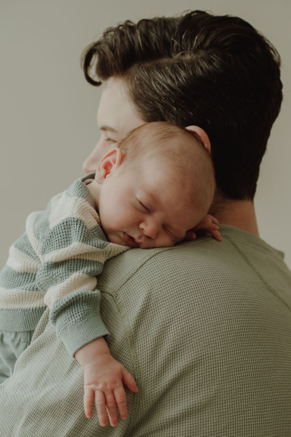 A father gently holds his sleeping newborn baby against his shoulder in a tender embrace.
