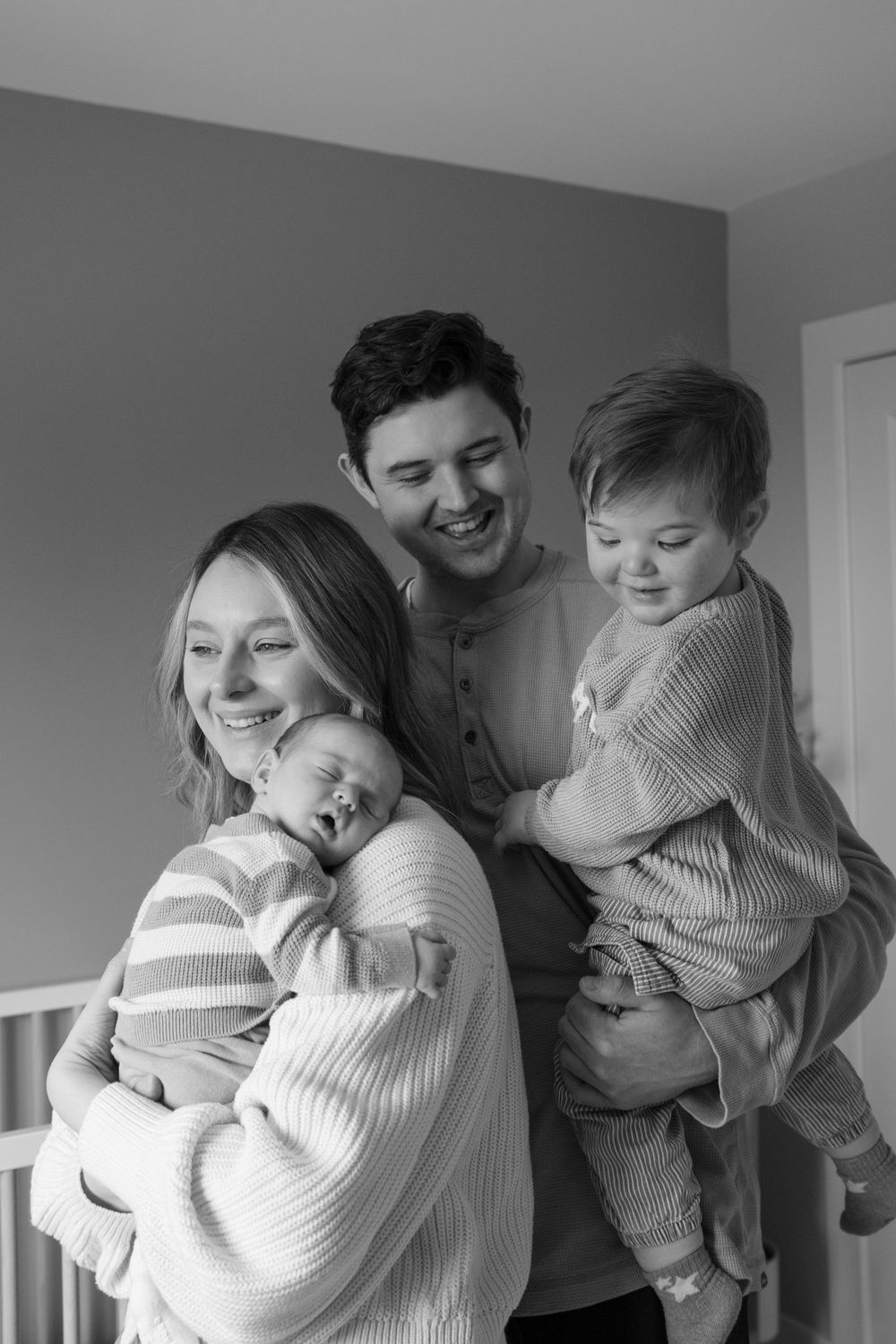 Black and white family portrait of smiling parents holding their newborn and toddler child indoors.