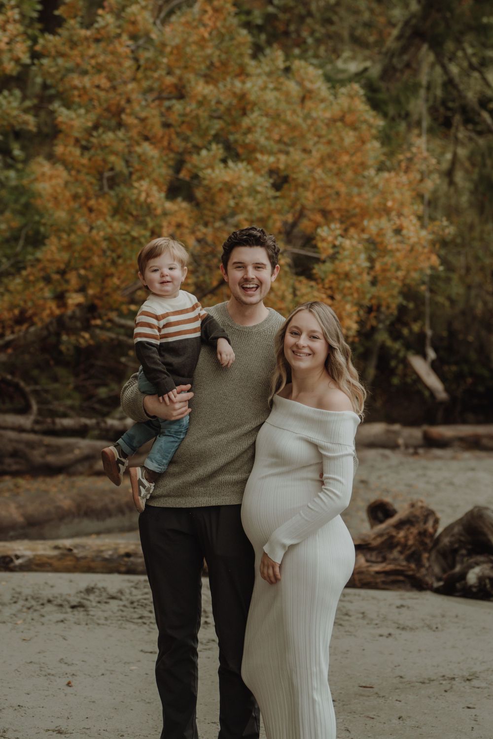 Smiling family maternity photo with toddler and pregnant mother in white dress on autumn beach.