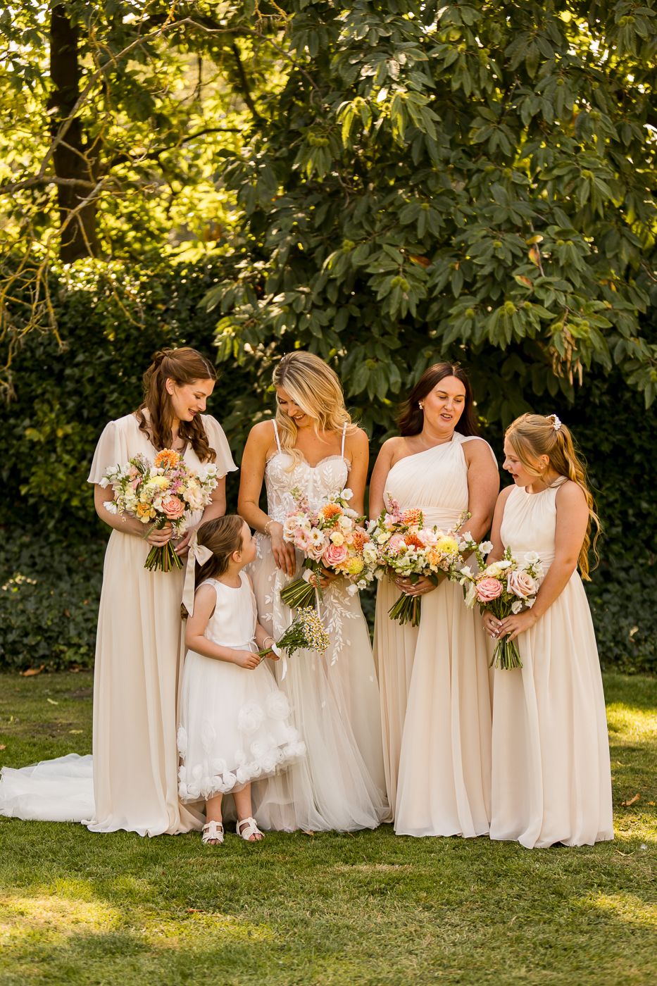 Wedding party wearing white and cream dresses poses together on grass with floral bouquets in a garden setting.
