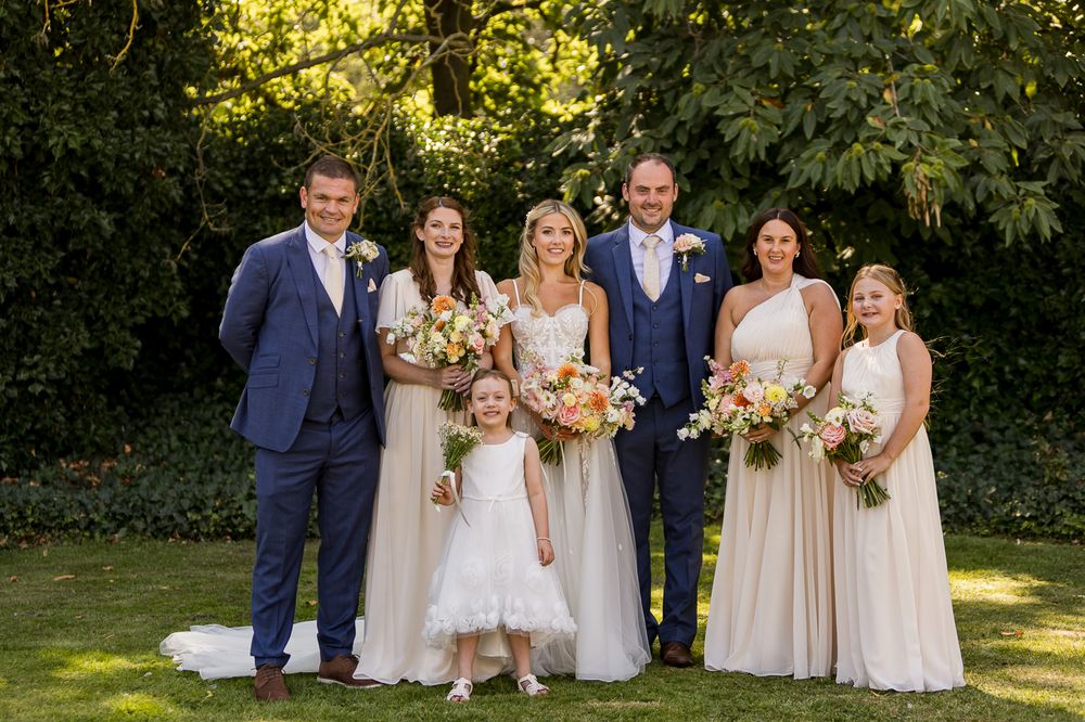 Wedding party poses in a garden setting with elegant attire and floral bouquets in natural sunlight.