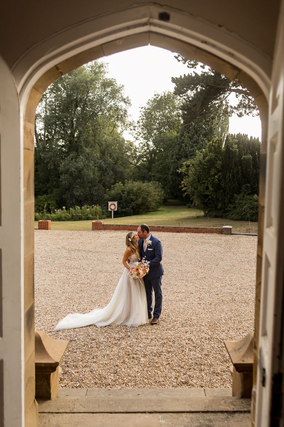 A couple in wedding attire embraces on a gravel courtyard, viewed through an elegant stone archway.