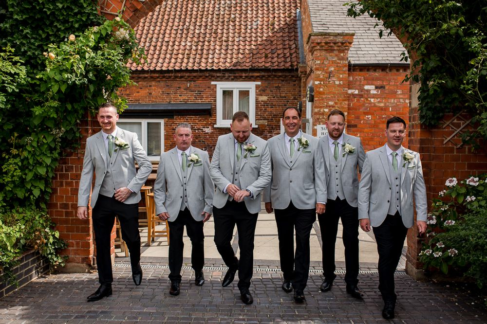 A group of groomsmen in matching light gray suits walk together on a path in front of a brick building.
