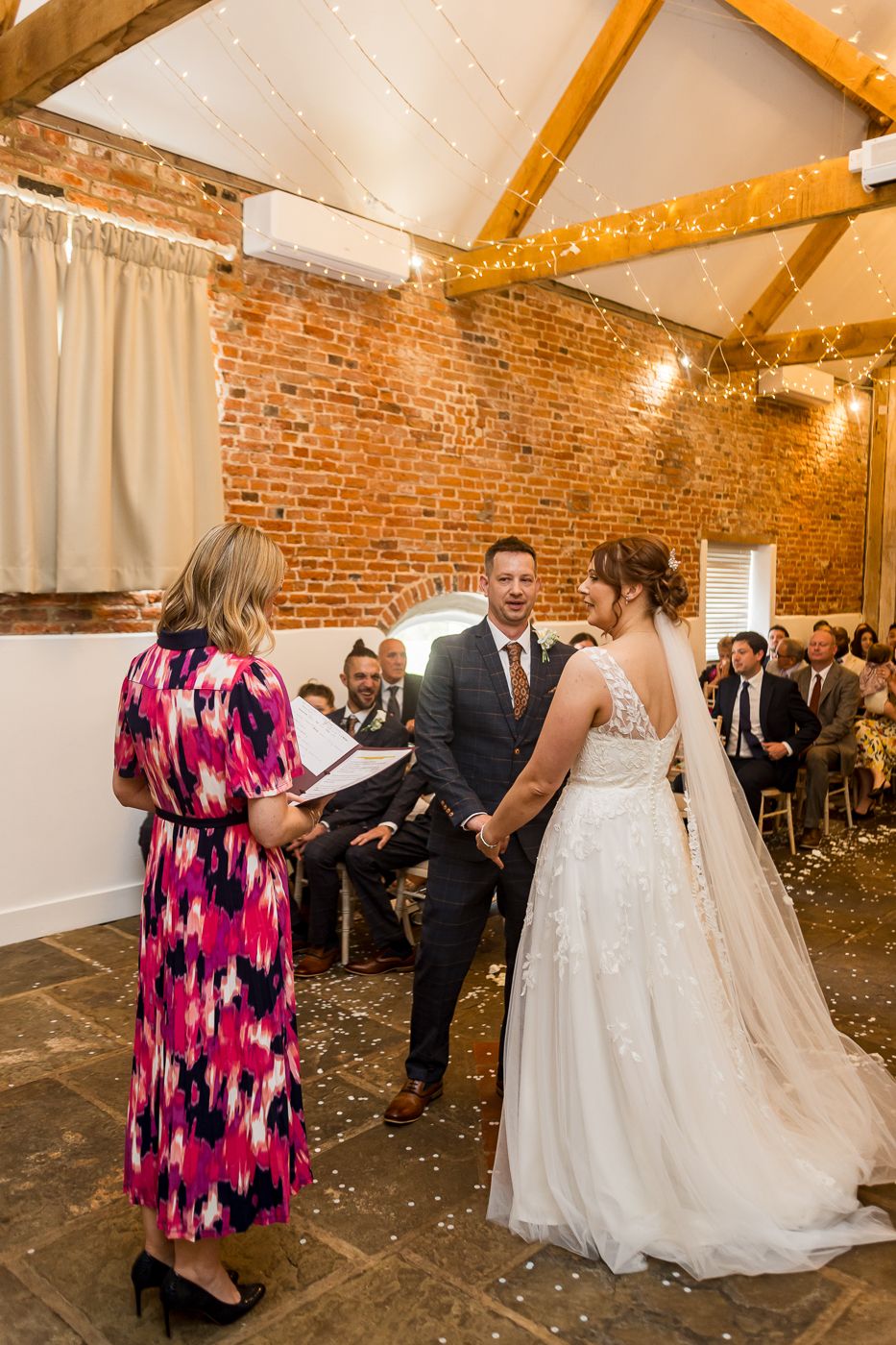 A wedding ceremony takes place in a rustic brick-walled venue with exposed wooden beams and string lights overhead.