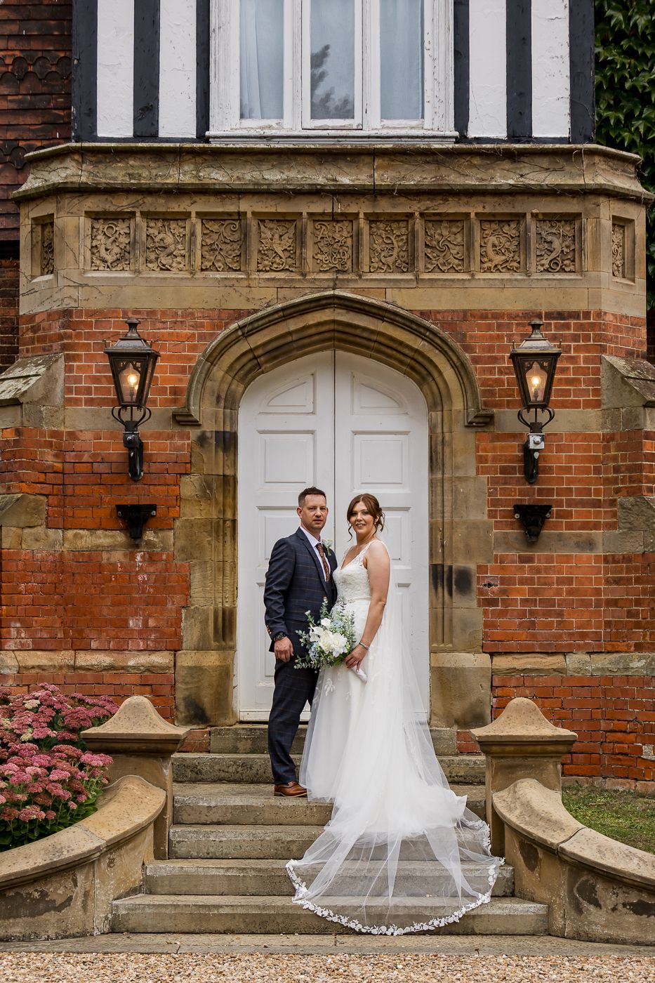 Wedding couple poses elegantly on historic brick building steps with ornate stone archway and blooming flowers.