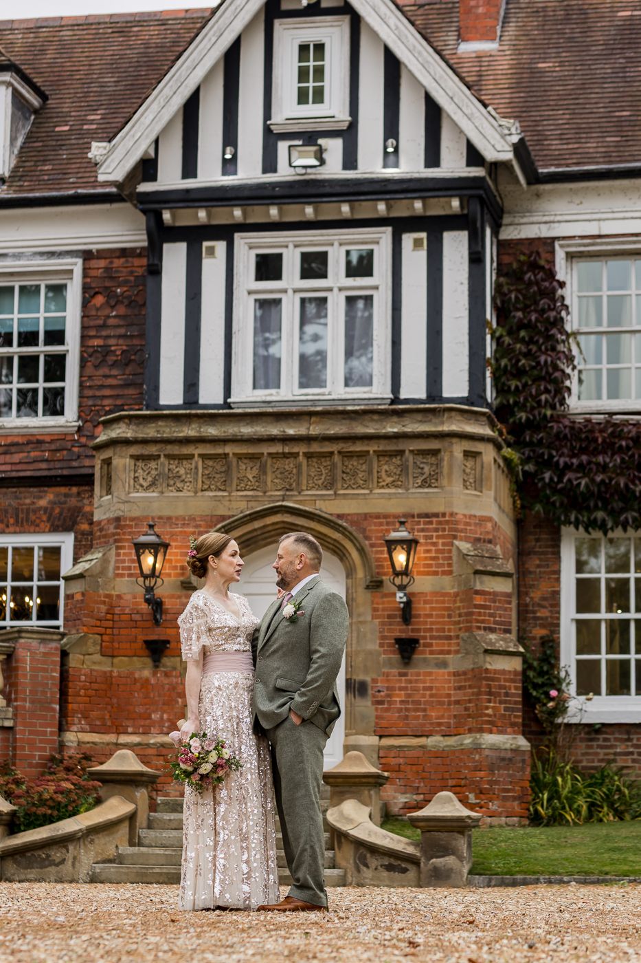 A couple shares romantic moments in front of a Tudor-style brick mansion with white trim and arched doorways.